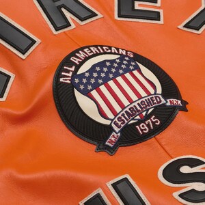 May include: Close-up of an orange leather jacket with black and white lettering and patches. The jacket features a circular patch with the text "ALL AMERICANS ESTABLISHED 1975 NY" and a shield design with the American flag.