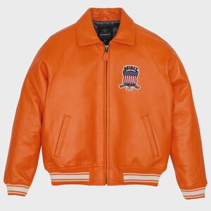 May include: Orange leather jacket with a bomber style. Features a collar, zipper closure, and a patch on the chest with a patriotic design. The jacket has white stripes on the cuffs and hem. The brand name "AVIREX" is visible.