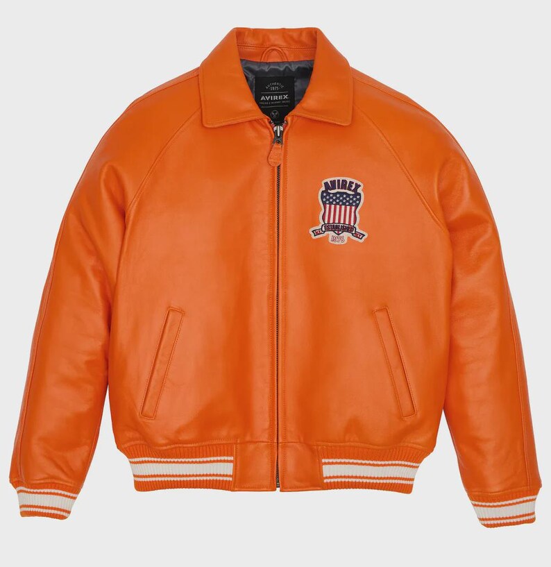 May include: Orange leather jacket with a bomber style. Features a collar, zipper closure, and a patch on the chest with a patriotic design. The jacket has white stripes on the cuffs and hem. The brand name "AVIREX" is visible.