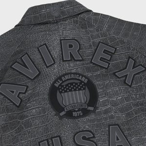 Puede incluir: Primer plano de una chaqueta gris con efecto piel de cocodrilo. La chaqueta presenta la palabra "AVIREX" en letras en relieve, junto con un parche circular que dice "ALL AMERICANS ESTABLISHED 1975". Un peque&ntilde;o emblema est&aacute; en el cuello.