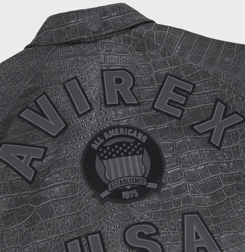 Puede incluir: Primer plano de una chaqueta gris con efecto piel de cocodrilo. La chaqueta presenta la palabra "AVIREX" en letras en relieve, junto con un parche circular que dice "ALL AMERICANS ESTABLISHED 1975". Un peque&ntilde;o emblema est&aacute; en el cuello.