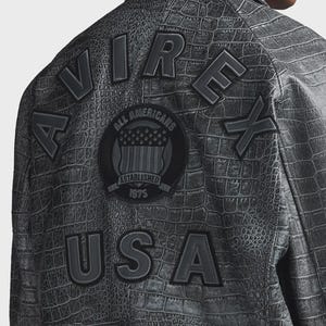 Puede incluir: Chaqueta gris con textura de piel de cocodrilo. La parte trasera de la chaqueta presenta las palabras "AVIREX USA" en letras grandes en relieve. Un parche circular dice "ALL AMERICANS ESTABLISHED 1975".