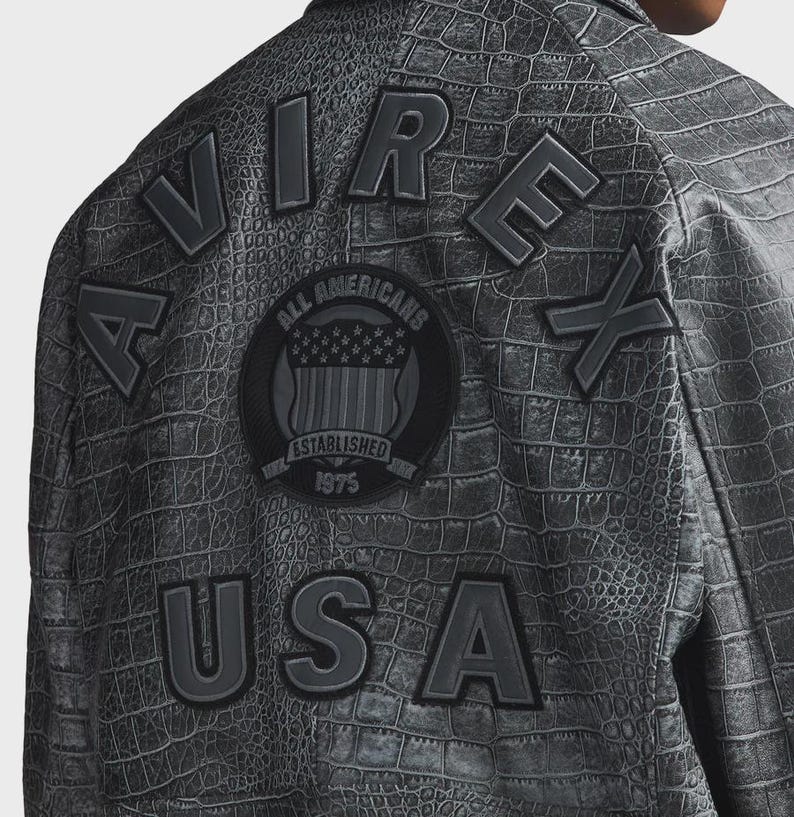 Puede incluir: Chaqueta gris con textura de piel de cocodrilo. La parte trasera de la chaqueta presenta las palabras "AVIREX USA" en letras grandes en relieve. Un parche circular dice "ALL AMERICANS ESTABLISHED 1975".