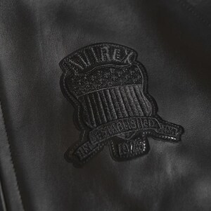 Puede incluir: Primer plano de una chaqueta de cuero negra con un parche texturizado. El parche tiene forma de escudo y muestra la marca "AVIREX" y el texto "ESTABLISHED NY 1975". La chaqueta tiene un ligero brillo.