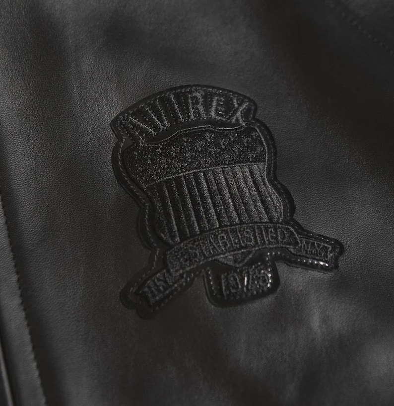 Puede incluir: Primer plano de una chaqueta de cuero negra con un parche texturizado. El parche tiene forma de escudo y muestra la marca "AVIREX" y el texto "ESTABLISHED NY 1975". La chaqueta tiene un ligero brillo.