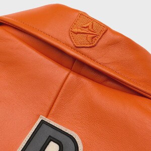 May include: Close-up of an orange leather jacket with a stitched emblem on the upper right shoulder. The jacket features a large letter patch with a black outline and a cream-colored border. The leather has a smooth, glossy finish.