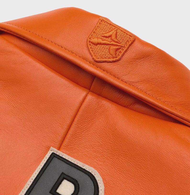 May include: Close-up of an orange leather jacket with a stitched emblem on the upper right shoulder. The jacket features a large letter patch with a black outline and a cream-colored border. The leather has a smooth, glossy finish.