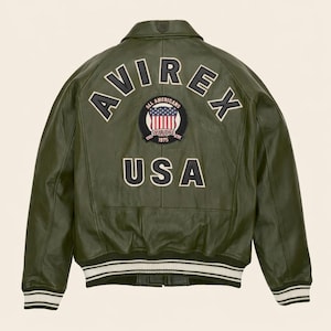 May include: Olive green leather jacket with the text "AVIREX" across the back, along with a patch featuring the American flag and the text "ALL AMERICANS ESTABLISHED 1975 USA". The jacket has a ribbed collar and cuffs with white stripes.