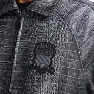 Puede incluir: Chaqueta gris texturizada con un patr&oacute;n de piel de cocodrilo. La chaqueta tiene cierre de cremallera y un parche negro en el pecho con el texto "AVIREX ESTABLISHED 1975" y un dise&ntilde;o de escudo con estrellas y rayas.
