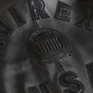 Puede incluir: Primer plano de una chaqueta de cuero negra con letras y parches en relieve. La chaqueta presenta la palabra "IREX" y un parche circular con el texto "All American" y "Established 1975". La chaqueta tiene una est&eacute;tica cl&aacute;sica y vintage.