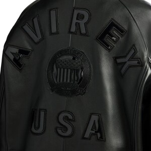 Puede incluir: Chaqueta de cuero negra con la palabra "AVIREX" en letras negras en relieve en la espalda. Tambi&eacute;n es visible un emblema circular con las palabras "ALL AMERICANS" y "USA". La chaqueta tiene un estilo bomber cl&aacute;sico.