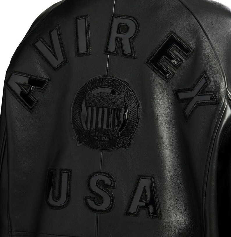 Puede incluir: Chaqueta de cuero negra con la palabra "AVIREX" en letras negras en relieve en la espalda. Tambi&eacute;n es visible un emblema circular con las palabras "ALL AMERICANS" y "USA". La chaqueta tiene un estilo bomber cl&aacute;sico.