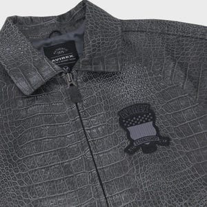 Puede incluir: Chaqueta gris texturizada con un patr&oacute;n de piel de cocodrilo. La chaqueta tiene cierre de cremallera y un peque&ntilde;o parche en el pecho con el texto "AVIREX" y "ESTABLISHED". La chaqueta tiene cuello y el texto "AUTHENTIC 1975" en la etiqueta.