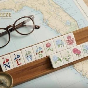 May include: A wooden tile holder with decorative tiles featuring floral and nautical designs. The tiles spell out "NEWS" and display various flower and sailboat illustrations. A pair of brown glasses and a compass are also present.