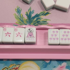 K&ouml;nnte beinhalten: Ein rosa Mahjong-Rack zeigt wei&szlig;e Spielsteine mit verschiedenen Designs. Einige Spielsteine zeigen einen rosa Hund, andere die Zahl sechs in Rosa und wieder andere rosa Blumen. Das Rack befindet sich vor einem bunten floralen Hintergrund.