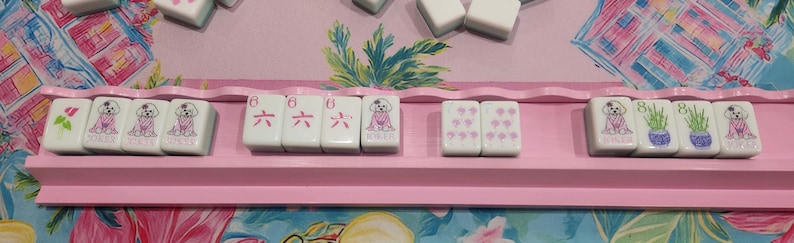 K&ouml;nnte beinhalten: Ein rosa Mahjong-Rack zeigt wei&szlig;e Spielsteine mit verschiedenen Designs. Einige Spielsteine zeigen einen rosa Hund, andere die Zahl sechs in Rosa und wieder andere rosa Blumen. Das Rack befindet sich vor einem bunten floralen Hintergrund.