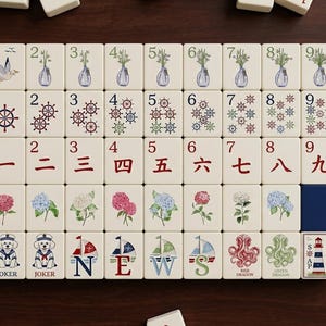May include: A complete set of ivory-colored Mahjong tiles. The tiles feature various nautical and floral designs, including sailboats, anchors, and flowers. Numbers and Chinese characters are also present on the tiles.