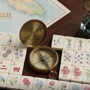 May include: Cream-colored tiles with floral and nautical illustrations, numbered 1-9, and Chinese characters. A vintage brass compass and inkwell with a quill sit nearby. The text "GULF BREEZE TILES" is visible.