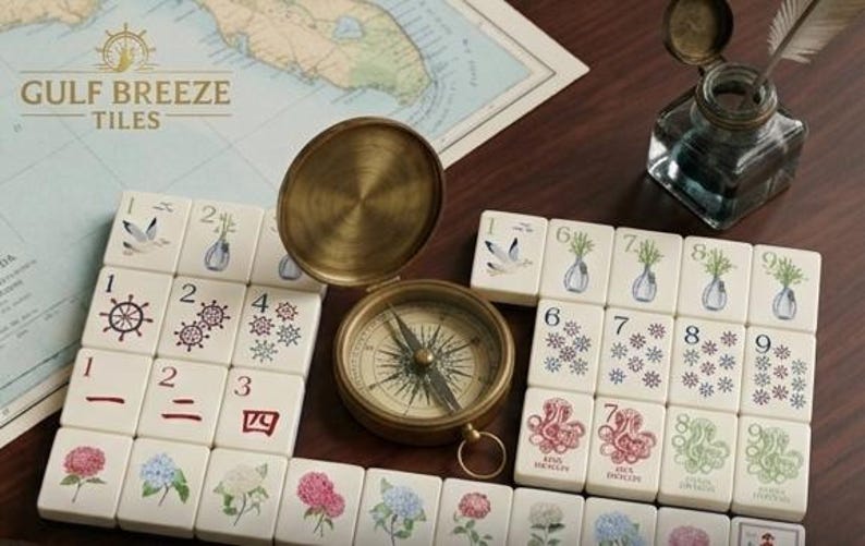 May include: Cream-colored tiles with floral and nautical illustrations, numbered 1-9, and Chinese characters. A vintage brass compass and inkwell with a quill sit nearby. The text "GULF BREEZE TILES" is visible.
