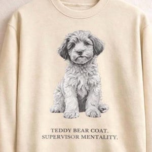 May include: A cream-coloured sweatshirt with a detailed grayscale illustration of a fluffy puppy. The puppy is sitting, with the text "TEDDY BEAR COAT. SUPERVISOR MENTALITY." printed below. The sweatshirt has long sleeves and a crew neck.
