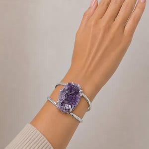 May include: A silver-toned cuff bracelet featuring a large, raw, purple amethyst druzy stone centerpiece. The bracelet has a split-shank design with two polished metal bands that hold the textured, crystalline gemstone securely in place.