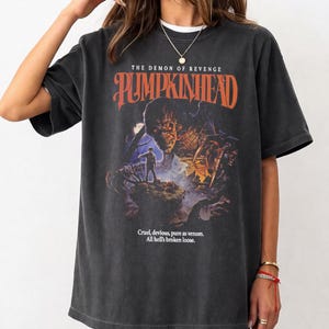 Può includere: T-shirt grigio scuro con il titolo del film "Pumpkinhead" in arancione. La grafica raffigura un mostro e un uomo in una scena oscura e nebbiosa. Il testo "Cruel, devious, pure as venom. All hell's broken loose" è stampato sotto.