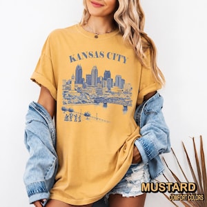 May include: Mustard-colored t-shirt with a blue graphic of the Kansas City skyline. The text "KANSAS CITY" is above the skyline. The shirt has rolled-up sleeves and is paired with a denim jacket.