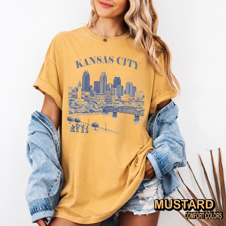 May include: Mustard-colored t-shirt with a blue graphic of the Kansas City skyline. The text "KANSAS CITY" is above the skyline. The shirt has rolled-up sleeves and is paired with a denim jacket.