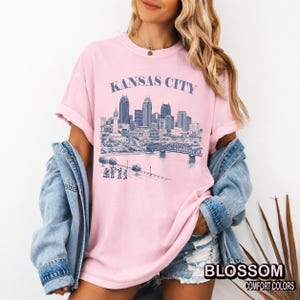 May include: A light pink t-shirt with the words "KANSAS CITY" above a blue and white illustration of a city skyline. The shirt also has the words "BLOSSOM COMFORT COLORS" printed on the bottom right.