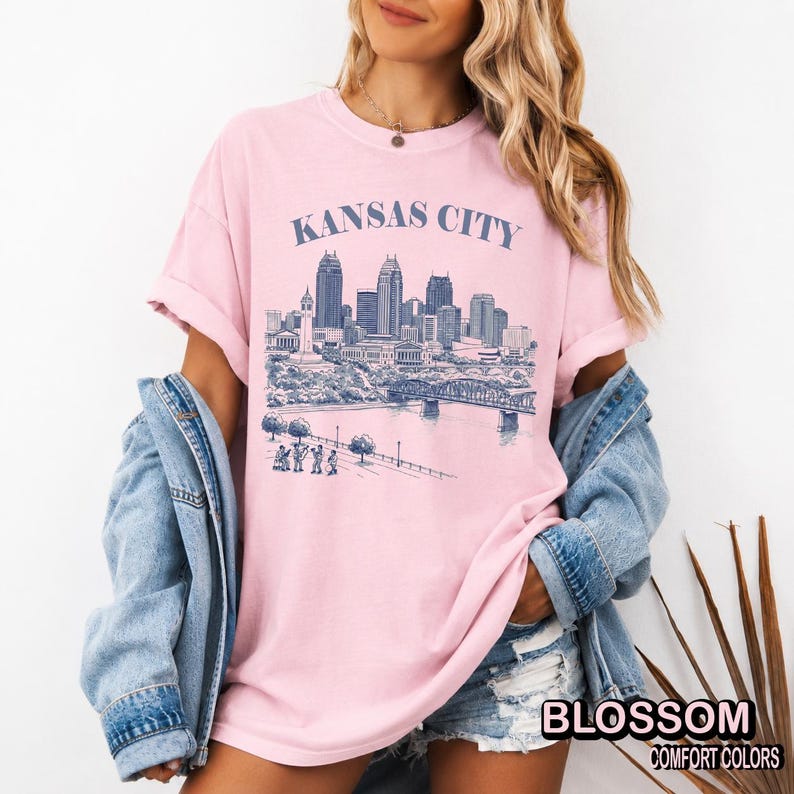 May include: A light pink t-shirt with the words "KANSAS CITY" above a blue and white illustration of a city skyline. The shirt also has the words "BLOSSOM COMFORT COLORS" printed on the bottom right.