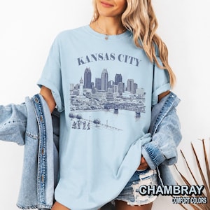 May include: Light blue t-shirt with the text "KANSAS CITY" above a detailed illustration of the city's skyline, including buildings, a bridge, and figures. The shirt also has the text "CHAMBRAY COMFORT COLORS".