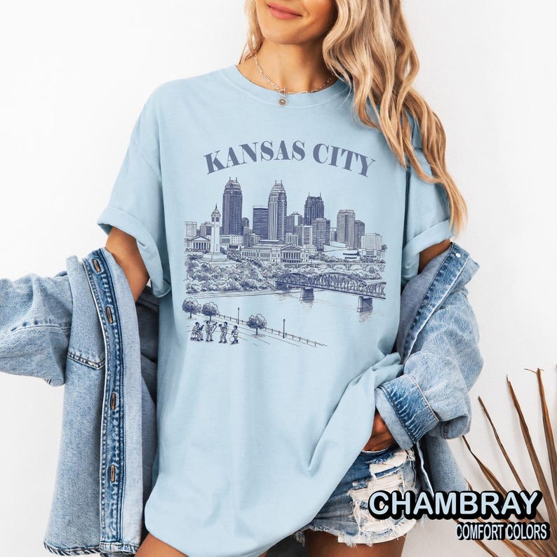 May include: Light blue t-shirt with the text "KANSAS CITY" above a detailed illustration of the city's skyline, including buildings, a bridge, and figures. The shirt also has the text "CHAMBRAY COMFORT COLORS".