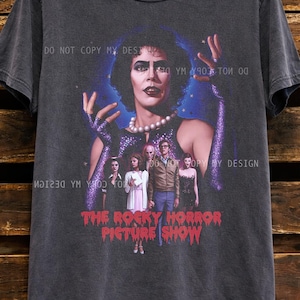 Può includere: T-shirt grigio scuro con grafica di "The Rocky Horror Picture Show". Il design include un ritratto di un personaggio con collana di perle e un gruppo di persone. Il testo "THE ROCKY HORROR PICTURE SHOW" è rosso.
