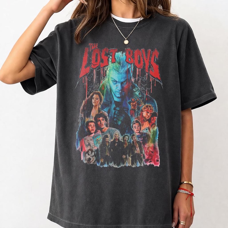 Pu&ograve; includere: T-shirt grigio scuro con una grafica colorata del film "The Lost Boys". Il design include il titolo del film in rosso e un collage di personaggi. La maglietta ha un aspetto vintage e usurato.