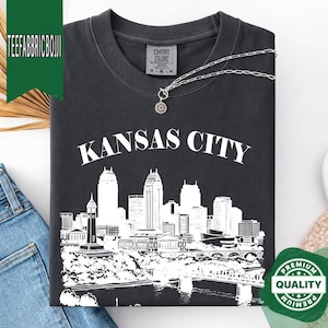 May include: Dark gray t-shirt with a white graphic of the Kansas City skyline and the city name. The shirt has a crew neck and short sleeves. A silver necklace with a pendant is draped on the shirt. The shirt is folded.