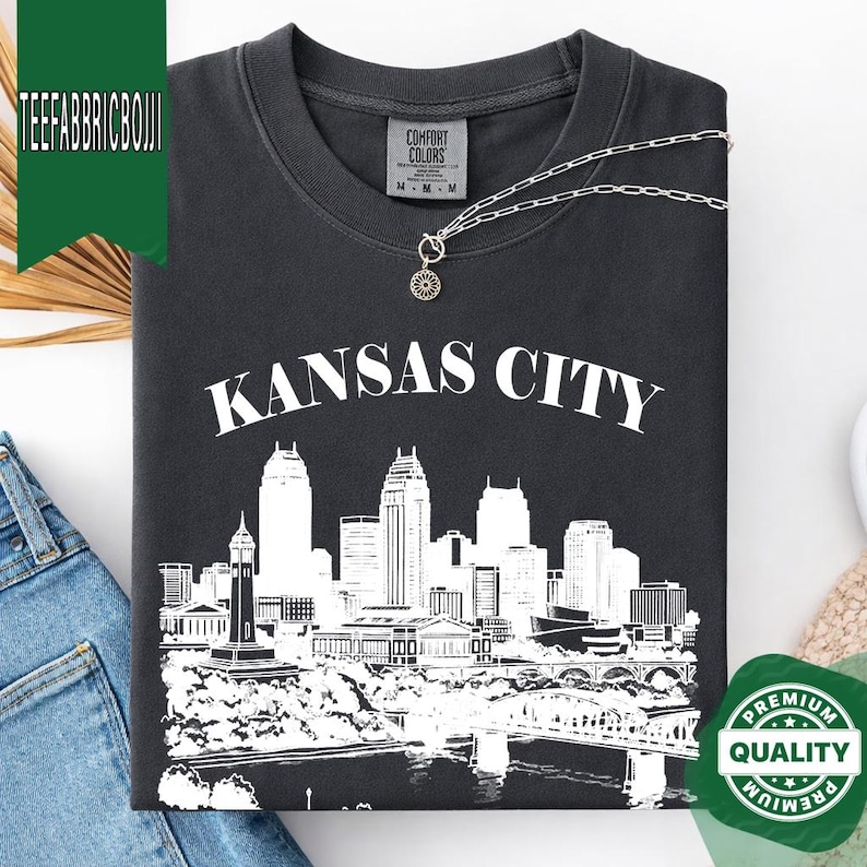 May include: Dark gray t-shirt with a white graphic of the Kansas City skyline and the city name. The shirt has a crew neck and short sleeves. A silver necklace with a pendant is draped on the shirt. The shirt is folded.