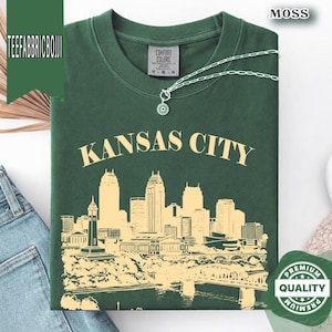 May include: A moss green t-shirt with "Kansas City" printed in a cream color above a cream-colored skyline graphic. The shirt has a crew neck and a small tag that says "Comfort Colors". A silver necklace with a pendant is on the shirt.