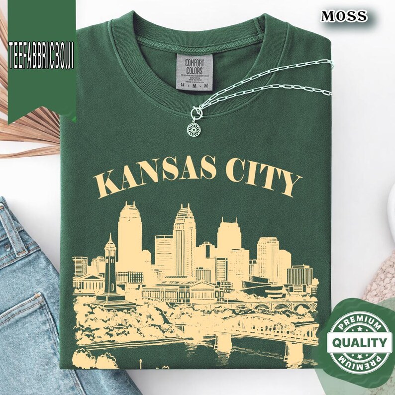 May include: A moss green t-shirt with "Kansas City" printed in a cream color above a cream-colored skyline graphic. The shirt has a crew neck and a small tag that says "Comfort Colors". A silver necklace with a pendant is on the shirt.