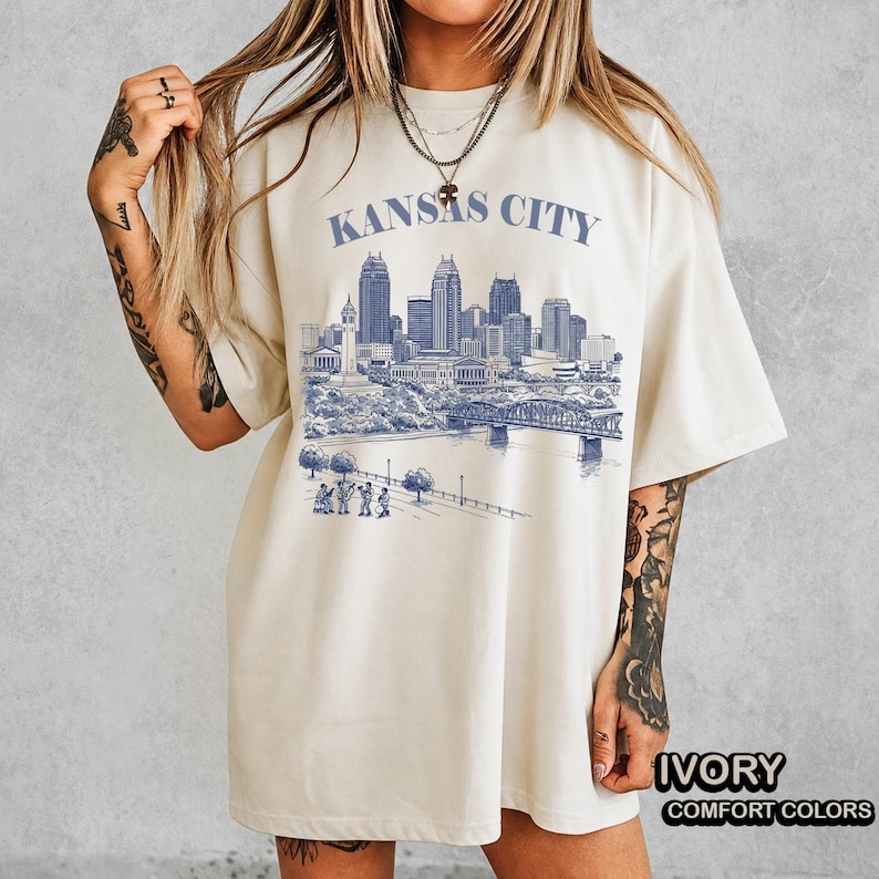 May include: Ivory-colored t-shirt featuring a blue illustration of the Kansas City skyline, including buildings, a bridge, and figures. The text "KANSAS CITY" is printed above the skyline. The shirt is labeled "IVORY" and "COMFORT COLORS".