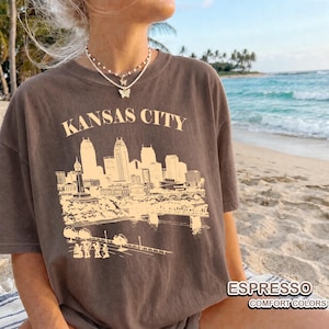 May include: Espresso-colored t-shirt featuring a beige graphic of the Kansas City skyline and the text "KANSAS CITY". The shirt also has the text "ESPRESSO" and "COMFORT COLORS".