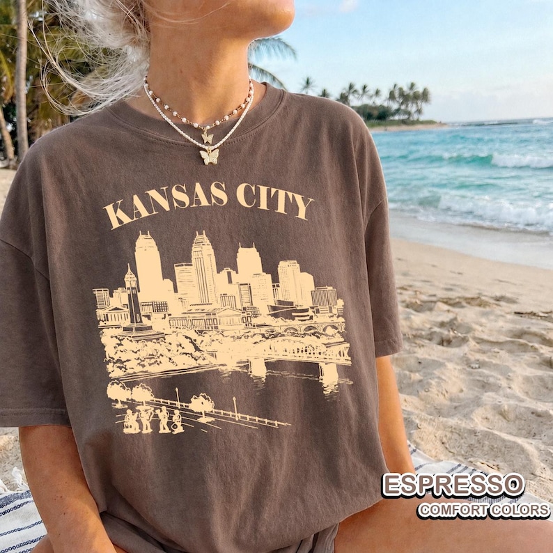 May include: Espresso-colored t-shirt featuring a beige graphic of the Kansas City skyline and the text "KANSAS CITY". The shirt also has the text "ESPRESSO" and "COMFORT COLORS".