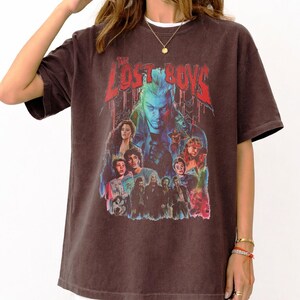 Pu&ograve; includere: T-shirt marrone con grafica del film "The Lost Boys". Il design include il titolo in rosso e un collage di personaggi nei toni del blu, rosso e viola. La maglietta ha maniche corte e girocollo.