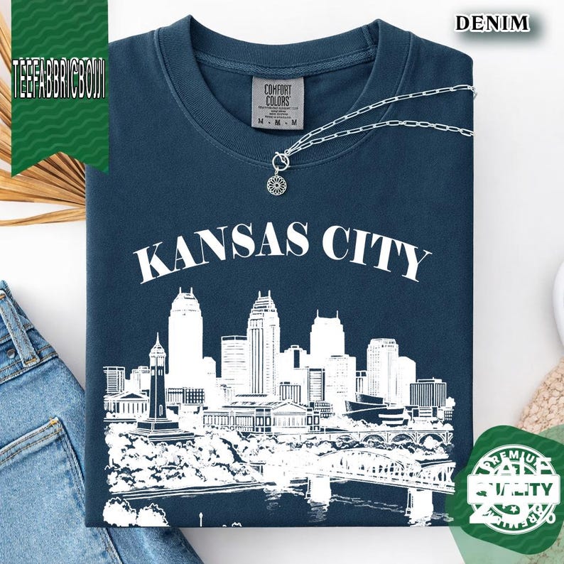 May include: A denim blue t-shirt with the words "Kansas City" in white block letters above a white skyline graphic. The shirt has a silver necklace with a small pendant. The shirt is labeled "Comfort Colors".