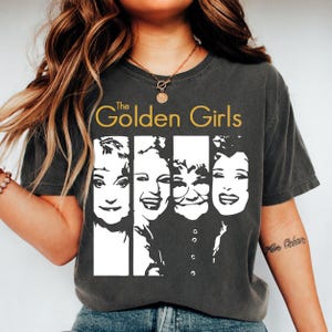 Può includere: T-shirt grigio scuro con la scritta dorata "The Golden Girls" sopra una grafica in bianco e nero dei quattro personaggi principali. La maglietta è a girocollo.