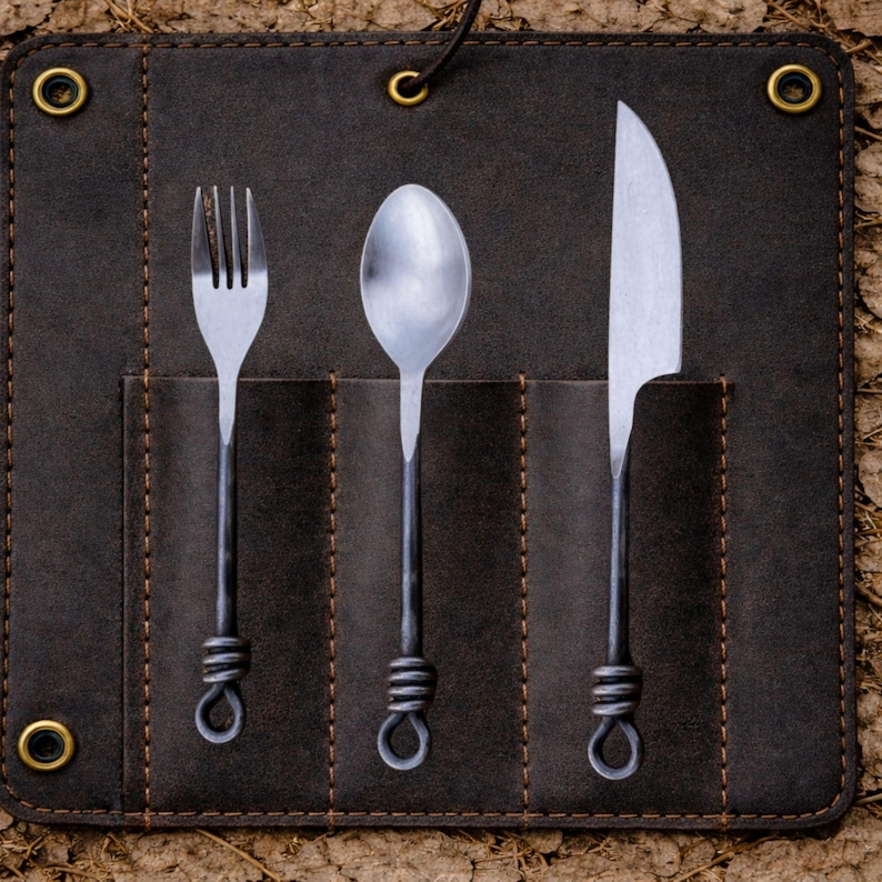 May include: A dark brown leather cutlery roll with a fork, spoon, and knife. The stainless steel utensils have dark metal handles with a looped design. The roll has brass eyelets and a leather tie closure, perfect for camping or outdoor dining.