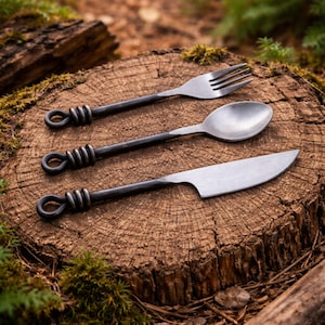 May include: Hand-forged, rustic cutlery set including a fork, spoon, and knife. The handles feature a unique coiled design, and the utensils are displayed on a weathered tree stump. The metal has a dark, matte finish, suggesting a handcrafted aesthetic.