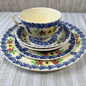 Peut inclure: Un ensemble tasse à thé et soucoupe en céramique empilé sur une assiette assortie. Les pièces de couleur crème présentent un motif décoratif bleu peint à la main avec de petites fleurs jaunes et rouges et des feuilles vertes en motif floral.