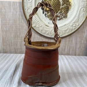 May include: A ceramic vessel with a reddish-brown glazed finish and a wide, flared rim. The container features a tall, arched handle constructed from twisted, natural brown vines that are securely attached to the sides of the pottery body.