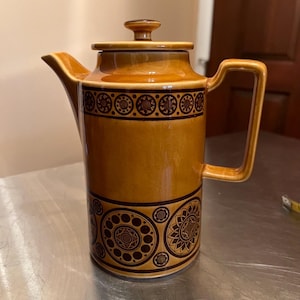 May include: A cylindrical ceramic coffee pot with a glossy mustard yellow glaze and dark brown geometric patterns. It features a matching lid with a small knob, a sharp angular spout, and a square-shaped handle. The body displays circular motifs.