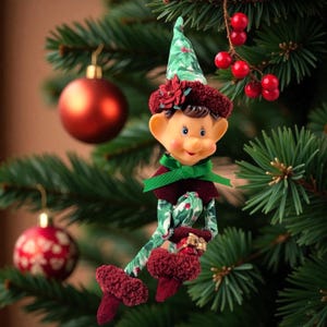 May include: A Christmas elf ornament featuring a green fabric hat and limbs with a holly leaf pattern, a maroon textured collar and shoes, a green ribbon bow at the neck, and a small gift box held in its hands, all accented by a red poinsettia.